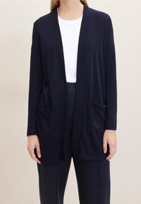Cardigan aperto navy realizzato in tessuto morbido, con maniche lunghe e due tasche frontali. Indossato sopra una maglietta bianca e pantaloni scuri.