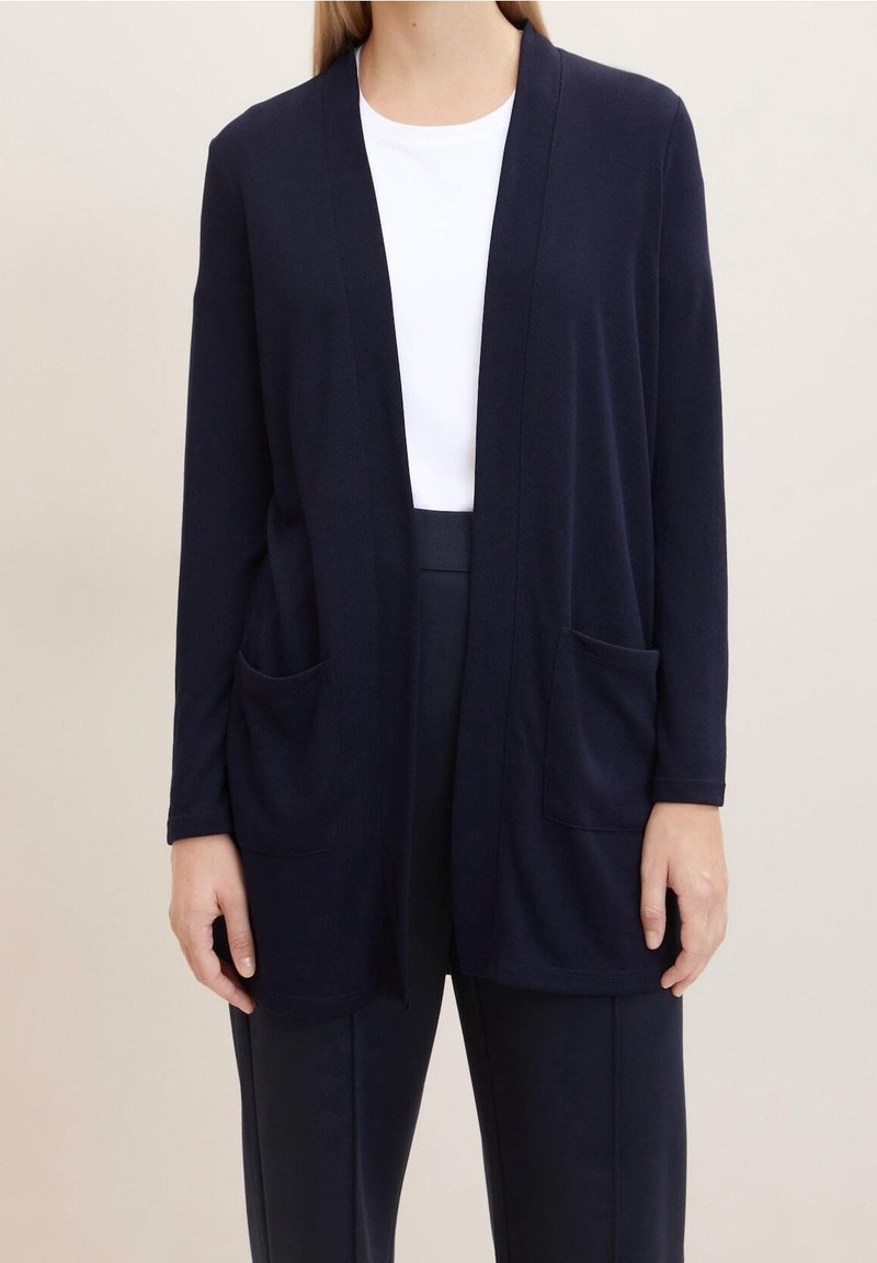 Cardigan aperto navy realizzato in tessuto morbido, con maniche lunghe e due tasche frontali. Indossato sopra una maglietta bianca e pantaloni scuri.