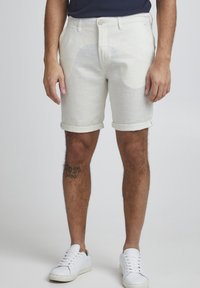 Pantalones cortos de algodón color beige claro con dobladillos enrollados, bolsillos frontales y un diseño de frente plano. Combinados con zapatillas blancas y con un tatuaje visible.