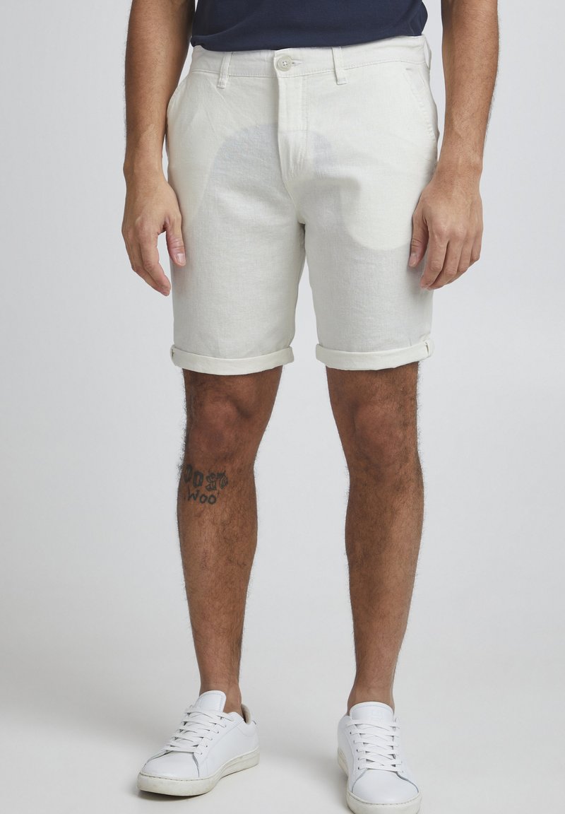 Pantalones cortos de algodón color beige claro con dobladillos enrollados, bolsillos frontales y un diseño de frente plano. Combinados con zapatillas blancas y con un tatuaje visible.