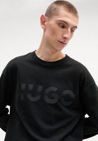 Juodas medvilnės džemperis su dideliu, toniniu "HUGO" logotipu priekyje. tekstūruota medžiaga ir apvalia iškirpte. Ilgos rankovės.