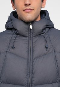 Giacca imbottita con colletto alto e cappuccio, realizzata in tessuto grigio scuro, dotata di zip frontale e cordini elastici per la regolazione.