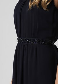 Robes bleu marine avec un design plissé, présentant une taille ornée de bijoux noirs qui ajoute de la texture et un détail visuel. Texture de tissu lisse.