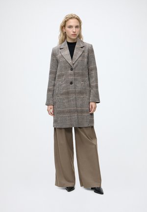 Femme portant un long manteau à carreaux marron sur un haut noir et un pantalon beige à jambes larges, debout devant un fond clair uni.