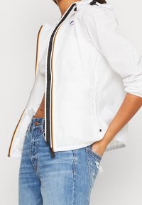 Witte lichtgewicht jas met een ritssluiting aan de voorkant, grijze accenten en oranje strepen. Voorzien van een capuchon en elastische manchetten, gedragen met een blauwe spijkerbroek.