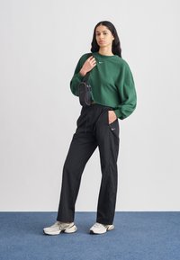 Πράσινο κοντό φούτερ με λευκό λογότυπο, μαύρο παντελόνι με ελαστική μέση και ανοιχτόχρωμα αθλητικά παπούτσια. Μικρή μαύρη τσάντα στερεωμένη στον ώμο.