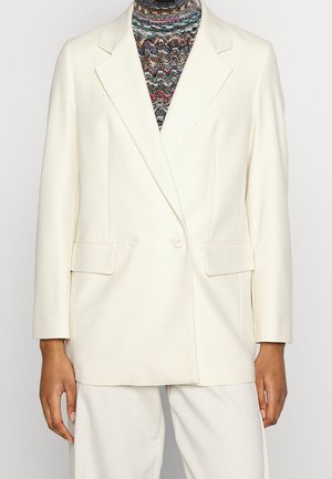 Personne portant un blazer croisé couleur crème sur un col roulé à motifs multicolores avec un pantalon crème assorti.