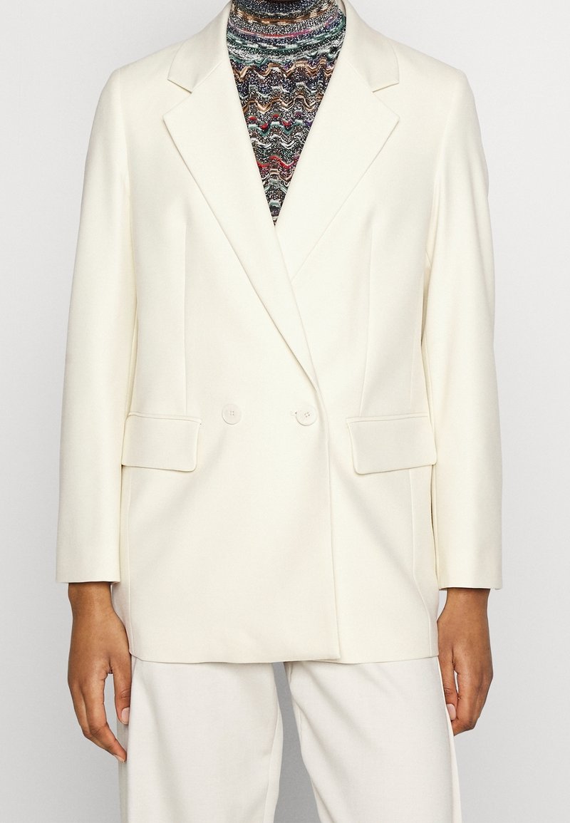 Personne portant un blazer croisé couleur crème sur un col roulé à motifs multicolores avec un pantalon crème assorti.
