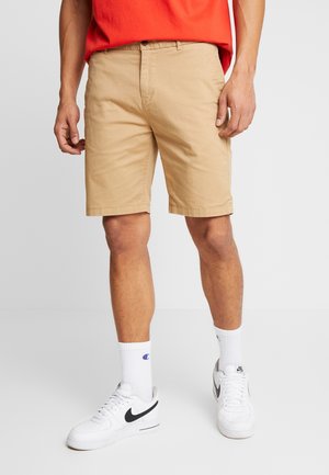 Mann trägt beige knielange Shorts, weiße Nike-Sneaker, weiße Champion-Socken und ein rotes Shirt, steht vor einfarbigem Hintergrund.