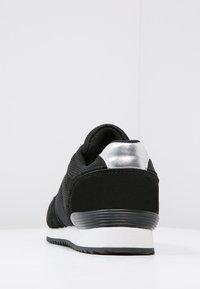 Chaussure de sport noire avec une tige texturée, accent de talon argenté et semelle extérieure blanche. Présente un bout arrondi et un design à lacets.