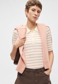 Πλεχτό πόλο με κοντά μανίκια σε κρεμ και ροζ ρίγες; συνδυασμένο με ένα ροζ πλεχτό πουλόβερ ριγμένο στους ώμους. Καφέ παντελόνι με κουμπί.