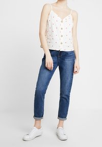 Mujer con una blusa blanca sin mangas abotonada con pequeñas flores rojas, jeans azules con puños y zapatillas blancas, de pie contra un fondo liso.