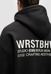 Sudadera con capucha negra que presenta texto impreso en blanco en la parte posterior, con una textura suave y un corte holgado, destacando los detalles de la marca.