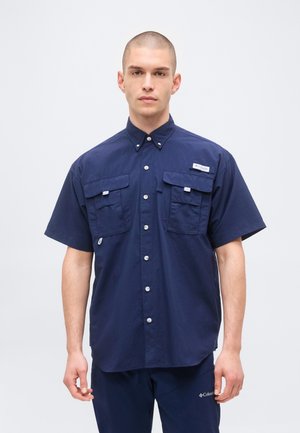 Homme debout face à l’avant portant une chemise bleu marine à manches courtes avec deux poches poitrine et un pantalon bleu marine assorti.