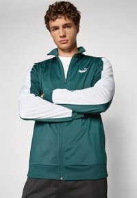 Tyrkis zip-jakke med hvite ermer, laget av lett stoff. Har PUMA-logo på brystet og elastiske mansjetter ved håndleddene.