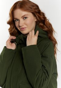 Chaqueta con capucha de color verde oliva, de textura suave, con cuello ajustable y cremallera frontal. Presenta un sutil logotipo en la manga y un corte relajado.