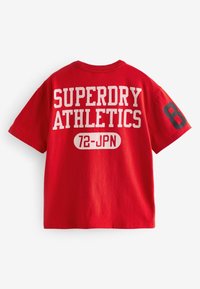 Rød bomulds-t-shirt med "SUPERDRY ATHLETICS" i hvid tekst og "72-JPN." Sort accent med tallet "8" på højre ærme.