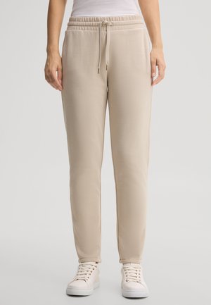 Unterkörper einer Person, die beige Hosen mit Kordelzug und weiße Sneakers trägt, vor einem hellgrauen Hintergrund stehend.