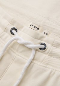 Nærbillede af beige snørebuks, der viser hvid reb-snor træde gennem sorte metaløjer og et stofmærke med "ombre" og størrelse M.