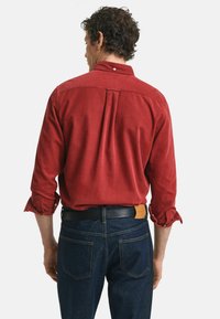 Chemise en velours côtelé rouge avec col boutonné, manches longues retroussées, détail de pli au dos ; portée avec un jean en denim foncé et une ceinture noire.