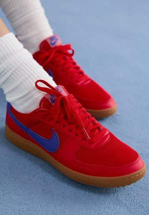 Fødder iført knaldrøde Nike-sneakers med blå swoosh-logoer og hvide sokker på en lyseblå struktureret gulv.