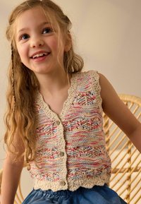 Cardigan senza maniche lavorato a maglia con motivi colorati in beige, rosa e blu, caratterizzato da un bordo scallop e chiusura a bottoni. Indossato su una gonna di jeans.
