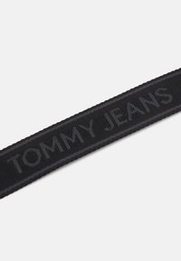 Etichetta in tessuto nero intrecciato con testo in rilievo "TOMMY JEANS" di colore grigio scuro. I bordi sono cuciti con precisione; senza ulteriori motivi.