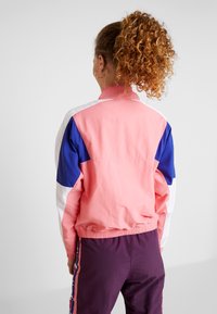 Chaqueta cortavientos colorida en rosa, azul y blanco, con un corte relajado, dobladillo elástico y diseño con paneles en contraste. Detalle de tejido texturizado visible.