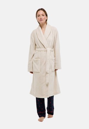 Femme debout pieds nus portant un peignoir beige noué à la taille sur un pantalon de pyjama bleu marine avec des motifs de cœurs rouges.