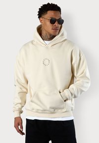 Cream-colored hoodie made from soft fabric, featuring a front pocket and circular graphic design on the chest. Includes a drawstring hood.