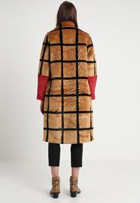 Mujer vista por detrás con un abrigo de piel a cuadros marrón con mangas rojas, pantalones negros cortos y botines de cuero de serpiente.