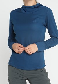 Långärmad, hooded tröja i mörkblått, gjord av texturerad tyg. Har en liten logotyp på bröstet och en rundad nederkant.