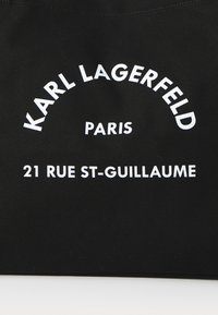 Sac fourre-tout noir avec texte blanc : "KARL LAGERFELD PARIS 21 RUE ST-GUILLAUME," avec une texture lisse et une forme rectangulaire.