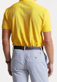 Osoba oblečená v žlutém polo tričku s krátkým rukávem, zasunutém do modro-bílých pruhovaných kalhot s multicolor pruhovaným opaskem, viděná zezadu.