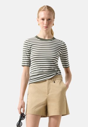 Kvinde iført grøn- og hvidstribet kortærmet skjorte og beige shorts, der holder sorte solbriller i den ene hånd.