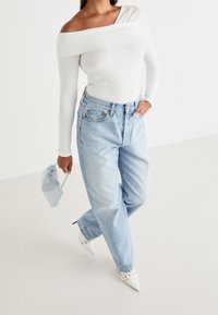 Žena v bílé topu s odhalenými rameny, světle modrých džínách s vysokým pasem, bílých špičatých lodičkách, drží malou třpytivou kabelku.