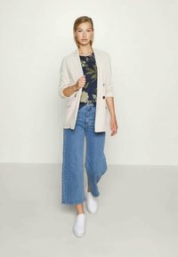 Blazer bege, blusa floral azul-marinho, jeans de perna larga azuis e ténis brancos. O blazer tem dois botões; a blusa possui padrões florais em verde e amarelo.