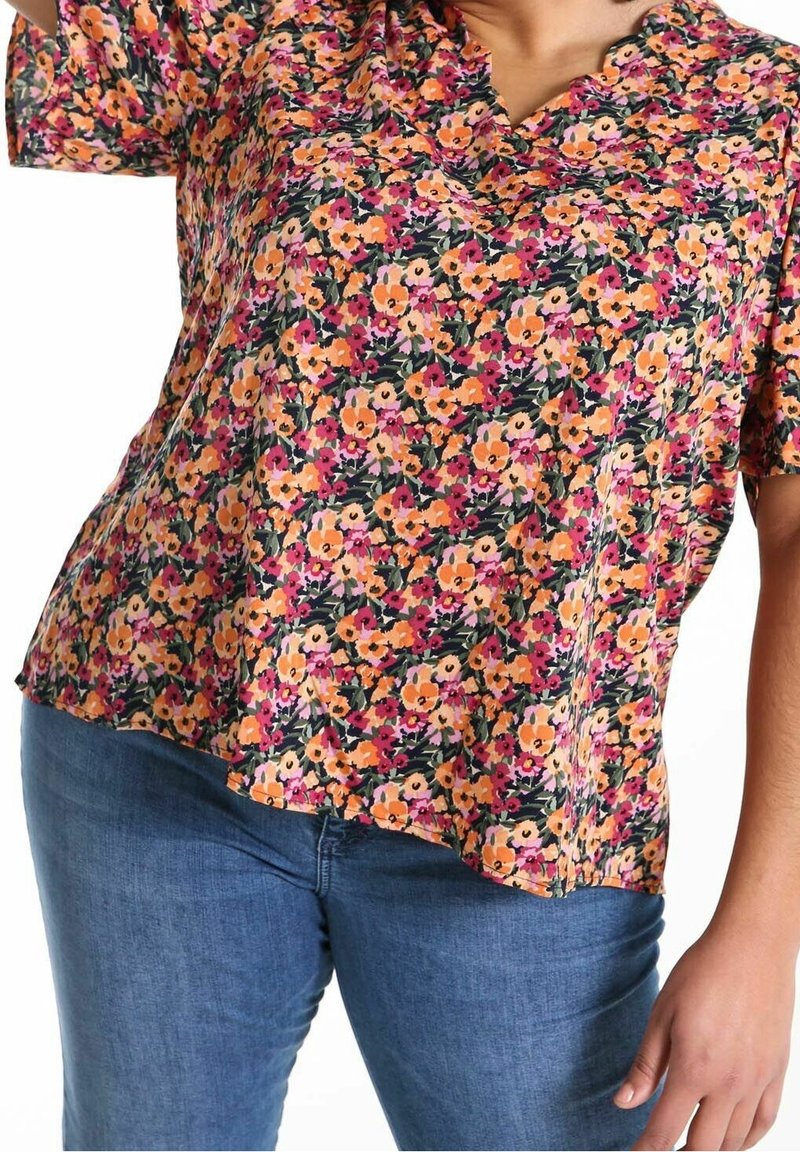 Persona che indossa una blusa a maniche corte colorata con fiori arancioni, rosa e viola abbinata a jeans blu.