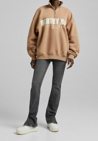 Sweatshirt zippé tan avec texte blanc, coupe décontractée, manches à revers, associé à un jean évasé gris foncé et des baskets blanches.