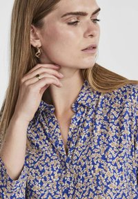 Lühikeste varrukatega sinine pluus, millel on beež lilleline muster, nööpidega ees ja kraega. Modell kannab kuldseid kõrvarõngaid ja sõrmust.