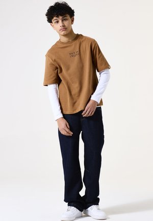 Bruine katoenen t-shirt met korte mouwen met de tekst "TERUG OVER 2 WEKEN" in het zwart, gedragen over een witte shirt met lange mouwen, gecombineerd met donkere spijkerbroek en witte schoenen.