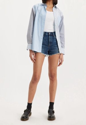 Camisa a rayas azul y blanca con botones, combinada con pantalones cortos de mezclilla desgastados. Zapatos negros con hebilla y calcetines oscuros completan el atuendo.