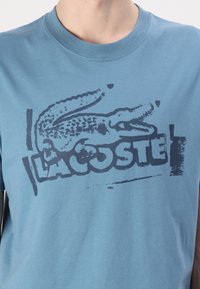 Jasnoniebieska bawełniana koszulka z krótkim rękawem, z okrągłym dekoltem i ciemną grafiką przedstawiającą krokodyla oraz napisem "LACOSTE," o gładkiej fakturze.