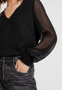 Svart blus med genomskinliga långärmar och V-ringning, med texturerade smockade manschetter; kombinerad med slitna grå denim.