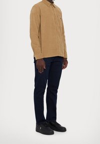 Uomo che indossa una camicia in velluto a coste color beige, jeans blu scuro e sneaker in pelle nera, in piedi su sfondo bianco.