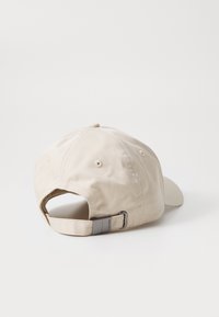 Beige bomullsbaseballcap med buet skjerm, justerbar stoffrem og metallisk detaljer bak. Glatt tekstur.