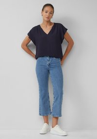 Blouse bleu marine à manches courtes et col en V portée avec un jean cropped bleu clair et des baskets blanches, sur un mannequin devant un arrière-plan neutre.