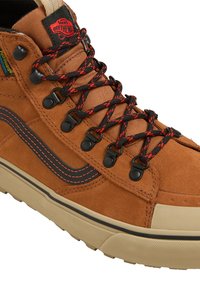 Brune semskede høyhælte sneakers med svarte gummidetaljer, røde og svarte snørebånd og en teksturert såle. Har en logoflagg på tungen.