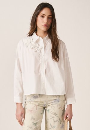 Frau mit langen braunen Haaren trägt ein locker sitzendes weißes Hemd mit Stoffblumen auf der Schulter und hell gemusterte, florale Hosen.