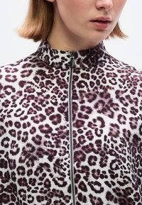 Τοπ με φερμουάρ σε λευκό και μωβ λεοπάρ μοτίβο, με ψηλό γιακά, υφή υφάσματος και λεπτομέρειες μαύρου φερμουάρ.